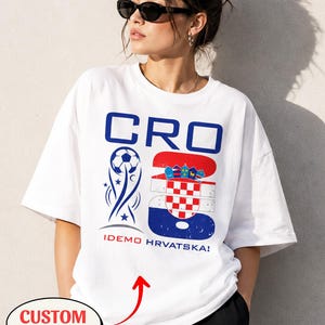 May include: A white short-sleeved t-shirt featuring a blue and red graphic design. The print includes the text CRO, a soccer ball icon, the number 26 with the Croatian flag pattern, and the text IDEMO HRVATSKA! in red and blue lettering.