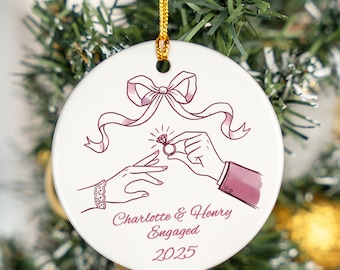 May include: White ceramic Christmas ornament with a pink illustration of a proposal scene. The design includes a bow, hands, and a ring, with the text "Charlotte & Henry Engaged 2025". A gold string is attached for hanging.