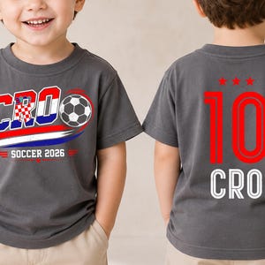 Może przedstawiać: Ciemnoszary t-shirt z grafiką z przodu przedstawiającą napis CRO, piłkę nożną oraz napis SOCCER 2026 w kolorach czerwonym, białym i niebieskim. Z tyłu koszulki znajduje się czerwona liczba 10 nad trzema czerwonymi gwiazdkami i białym napisem CRO.
