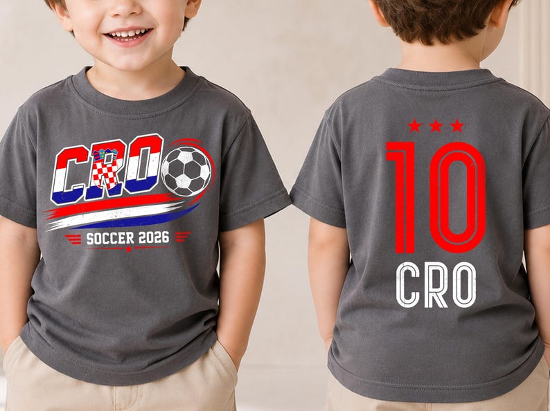 Może przedstawiać: Ciemnoszary t-shirt z grafiką z przodu przedstawiającą napis CRO, piłkę nożną oraz napis SOCCER 2026 w kolorach czerwonym, białym i niebieskim. Z tyłu koszulki znajduje się czerwona liczba 10 nad trzema czerwonymi gwiazdkami i białym napisem CRO.