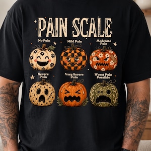 May include: A black t-shirt featuring a graphic titled PAIN SCALE with six pumpkins showing different expressions. The pumpkins are labeled No Pain, Mild Pain, Moderate Pain, Severe Pain, Very Severe Pain, and Worst Pain Possible with various patterns.