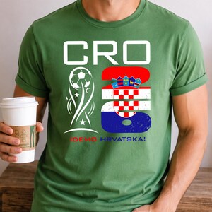 May include: A sage green short-sleeve t-shirt featuring a white graphic with the text CRO 26. The design includes a soccer ball icon and the Croatian flag pattern within the number 26, with the text IDEMO HRVATSKA! printed in red and blue below.