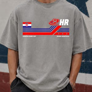 May include: A grey short-sleeve t-shirt featuring a graphic with the Croatian flag, a soccer ball, the letters HR, and the text CROATIA GAME DAY and IDEMO HRVATSKA! The design includes horizontal red and blue stripes and three small stars.