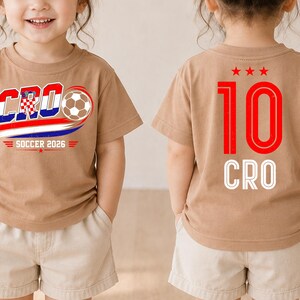 Może przedstawiać: Beżowy t-shirt z grafiką z przodu zawierającą napis CRO, piłkę nożną oraz SOCCER 2026. Z tyłu koszulki znajduje się czerwona liczba 10, trzy czerwone gwiazdki nad nią oraz biały napis CRO pod liczbą.