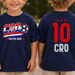 Może przedstawiać: Granatowa koszulka z grafiką z przodu zawierającą napis CRO, piłkę nożną oraz słowa SOCCER 2026. Z tyłu koszulki znajduje się czerwona liczba 10, trzy czerwone gwiazdki nad nią oraz napis CRO wykonany białymi literami pod numerem.