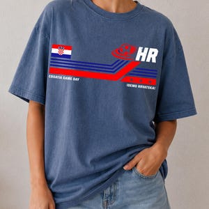 May include: A blue t-shirt featuring a graphic design with the Croatian flag, a soccer ball icon, and the text "CROATIA GAME DAY" and "IDEMO HRVATSKA!" alongside blue and red horizontal stripes. The shirt has a crew neckline and short sleeves.