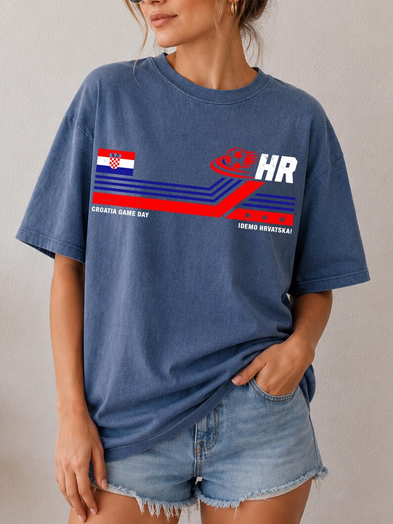 May include: A blue t-shirt featuring a graphic design with the Croatian flag, a soccer ball icon, and the text "CROATIA GAME DAY" and "IDEMO HRVATSKA!" alongside blue and red horizontal stripes. The shirt has a crew neckline and short sleeves.