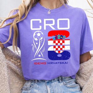May include: A purple short-sleeve t-shirt featuring the white text CRO and the number 26. The number 26 is filled with the Croatian flag pattern. A white soccer ball graphic and the red and white text IDEMO HRVATSKA! are printed below.