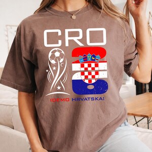 May include: A brown short-sleeve t-shirt featuring the white text CRO, a white soccer ball graphic, and a large number 23 filled with the red, white, and blue Croatian flag pattern. Below the graphics, the text IDEMO HRVATSKA! is printed in red and blue.