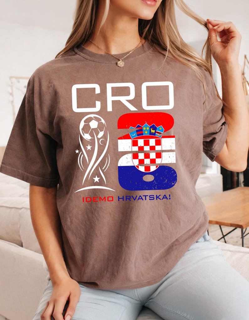 May include: A brown short-sleeve t-shirt featuring the white text CRO, a white soccer ball graphic, and a large number 23 filled with the red, white, and blue Croatian flag pattern. Below the graphics, the text IDEMO HRVATSKA! is printed in red and blue.