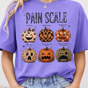 May include: A purple t-shirt featuring a graphic of six pumpkins arranged in two rows, labeled PAIN SCALE. Each pumpkin displays a different facial expression and pattern, representing levels from No Pain to Worst Pain Possible, with text labels.