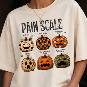 May include: A cream-colored t-shirt featuring a graphic titled PAIN SCALE with six illustrated pumpkins showing different facial expressions. The pumpkins are labeled No Pain, Mild Pain, Moderate Pain, Severe Pain, Very Severe Pain, and Worst Pain Possible.