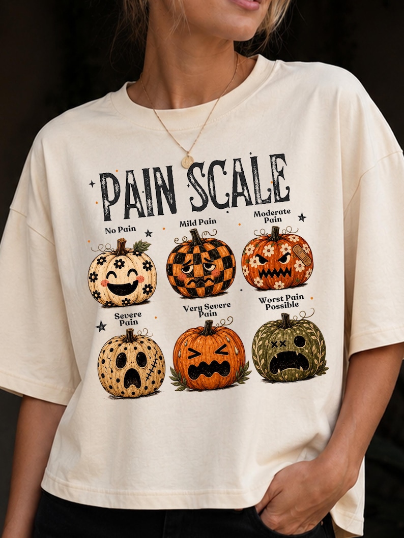 May include: A cream-colored t-shirt featuring a graphic titled PAIN SCALE with six illustrated pumpkins showing different facial expressions. The pumpkins are labeled No Pain, Mild Pain, Moderate Pain, Severe Pain, Very Severe Pain, and Worst Pain Possible.