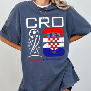 May include: A dark blue oversized t-shirt featuring a white graphic with the text CRO, a soccer ball icon, and the number 26 filled with the Croatian flag pattern. Below the graphic, the text IDEMO HRVATSKA! is printed in red and blue.