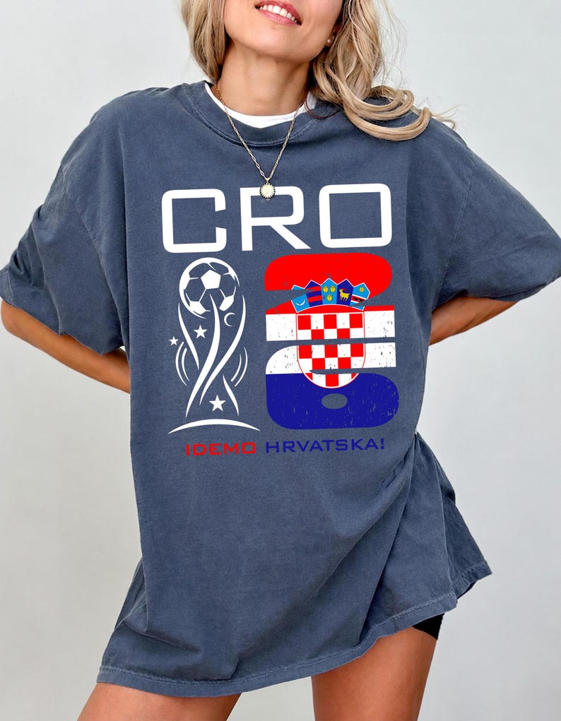 May include: A dark blue oversized t-shirt featuring a white graphic with the text CRO, a soccer ball icon, and the number 26 filled with the Croatian flag pattern. Below the graphic, the text IDEMO HRVATSKA! is printed in red and blue.