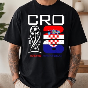 May include: A black t-shirt featuring a white graphic with the text CRO 26. The design includes a soccer ball trophy icon and the Croatian flag pattern within the number 26. Below the graphic, the text IDEMO HRVATSKA! is printed in red and blue.