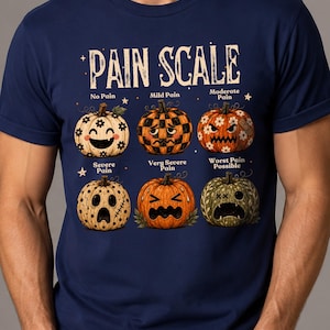 May include: A navy blue t-shirt featuring a graphic titled PAIN SCALE with six pumpkins showing different facial expressions. The pumpkins represent levels from No Pain to Worst Pain Possible, with various patterns, colors, and decorative elements.