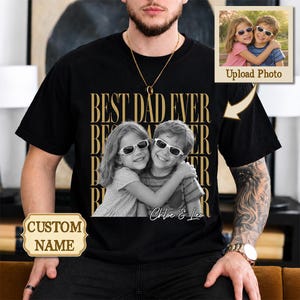 May include: A black t-shirt featuring a gold graphic print with the text BEST DAD EVER repeated in the background. A black and white photograph of two children wearing sunglasses is centered on the chest, with the names Chloe & Leo printed below.