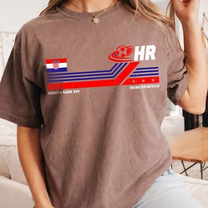 May include: A brown short-sleeve t-shirt featuring a graphic with the Croatian flag, a soccer ball, the text HR, the phrase CROATIA GAME DAY, and the text IDEMO HRVATSKA! with red and blue horizontal lines and three small red stars.