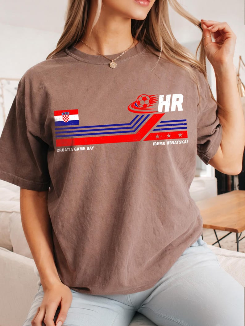 May include: A brown short-sleeve t-shirt featuring a graphic with the Croatian flag, a soccer ball, the text HR, the phrase CROATIA GAME DAY, and the text IDEMO HRVATSKA! with red and blue horizontal lines and three small red stars.