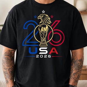 Può includere: Una t-shirt nera con un design grafico che presenta il numero 2026 in blu e rosso, un'aquila dorata e un trofeo dorato. Il testo USA 2026 è stampato sotto la grafica, accompagnato da tre piccole stelle nei colori blu, bianco e rosso.