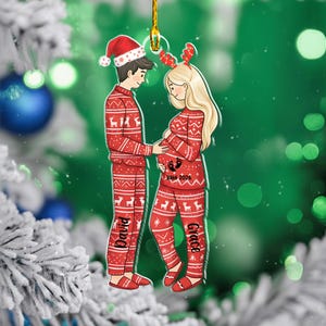 May include: A Christmas tree decoration depicting a couple in red festive pyjamas with white reindeer and snowflake designs. The woman is pregnant, and the ornament has the text "Baby 2026". The man wears a Santa hat, and the woman wears reindeer antlers. The names "David" and "Grace" are visible.