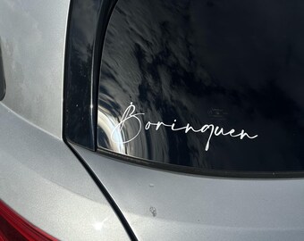 May include: A white vinyl decal featuring the word Borinquen in a cursive script font, applied to the rear window of a silver vehicle. The decal is positioned on the glass above the red taillight assembly, displaying a clean and simple design.