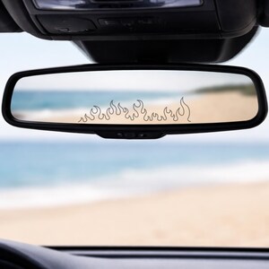 May include: A black-framed rectangular car rearview mirror featuring a thin, black line-art decal of stylized flames applied horizontally across the center of the glass, reflecting a blurred view of a sandy beach and the ocean horizon.