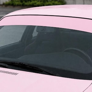 May include: A close-up of a pink car's windshield and roof. The windshield has a dark tint, and the car's body is a solid, light pink color. The wipers are visible at the base of the windshield.