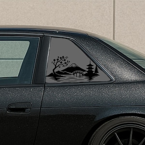 May include: A black vinyl decal applied to the rear side window of a dark car. The design features a silhouette of a mountain, a pagoda, a bridge, a cherry blossom tree, and water reflections, creating a detailed landscape scene on the glass.