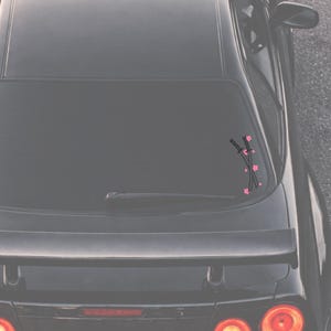 May include: Black car rear window with a vinyl decal featuring two crossed katanas and pink cherry blossoms. The design is a silhouette, with the swords and flowers in black and pink.