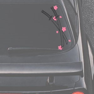 Pode incluir: Gráfico preto de duas katanas cruzadas com cabos detalhados e flores de cerejeira rosa. O design está na janela traseira de um veículo preto. A imagem inclui um limpador traseiro.