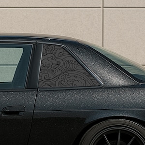 May include: A black car window with a decorative, dark gray wave pattern. The design is applied to the small, triangular window, adding a unique aesthetic to the vehicle's exterior. The car's body has a textured, dark gray finish.