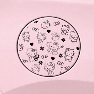 May include: A circular light pink car fuel door cover featuring a repeating black line art pattern of Hello Kitty characters, small hearts, and flowers. The design includes various poses of the character, including some with wings and strawberry hats.