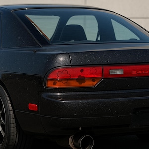 May include: Black sports car rear with cherry blossom tail light decals. The car has a 240SX emblem and a black exhaust pipe. The car's paint has a glittery finish.