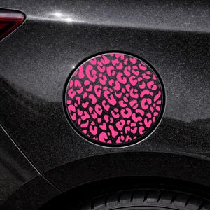 May include: A black car fuel door featuring a circular pink leopard print design. The design is a repeating pattern of irregular shapes. The car's body has a dark, glittery finish. A portion of the tyre is visible.