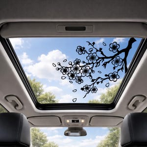 May include: A black cherry blossom branch decal is applied to the sunroof of a car, with a blue sky and clouds visible through the glass. The decal features detailed flower and leaf shapes, creating a decorative effect.