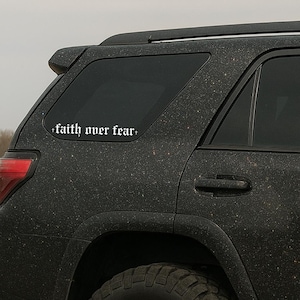 May include: A white vinyl decal with the phrase "faith over fear" in a gothic font is affixed to the rear window of a black SUV. The vehicle has a glittery, textured finish, and the decal is centered on the glass.