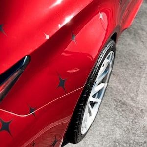 May include: Close-up of a red car's side panel with a star pattern. The car's wheel is visible, featuring a silver rim and black tire. The paint reflects light, highlighting the car's curves and design.