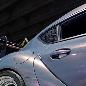 Könnte beinhalten: Ein metallic-grauer Sportwagen mit einem Aufkleber am hinteren Seitenfenster, der ein weißes Wellenmuster zeigt. Das Fahrzeug verfügt über einen Heckspoiler, einen Tankdeckel, einen Türgriff und einen Reifen mit der Aufschrift Extreme.
