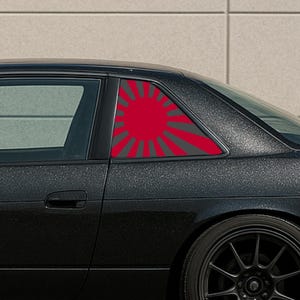 May include: A black car with a red and gray rising sun graphic on the rear window. The graphic is a stylized sunburst design. The car has black rims and a dark, textured paint finish.