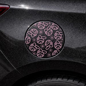 Könnte beinhalten: Ein schwarzer Tankdeckel mit einem rosa Hasen- und Blumenmuster. Der kreisförmige Deckel ist an einem schwarzen Auto angebracht, wobei ein Reifen unten sichtbar ist. Das Design zeigt Cartoon-Hasen und Blumen.