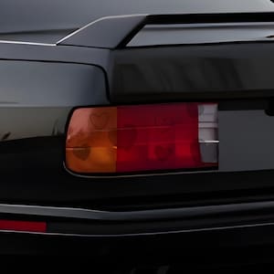 以下が含まれることがあります： 黒い車のリアエンドのクローズアップ。ハート型のデザインが施されたテールライトとスポイラーが特徴です。テールライトはオレンジと赤のセクションがあり、ハートがかすかに見えます。車のボディは光沢があり、周囲を反射しています。
