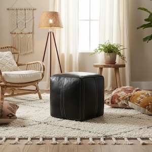 A modern black square Moroccan leather pouf ottoman with white stitching on a rug next to a bohemian rattan chair.
