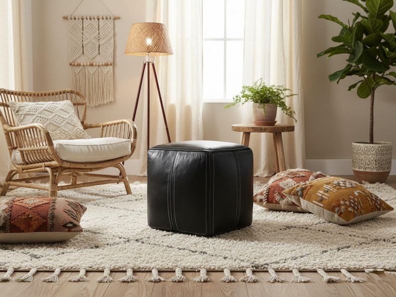 A modern black square Moroccan leather pouf ottoman with white stitching on a rug next to a bohemian rattan chair.