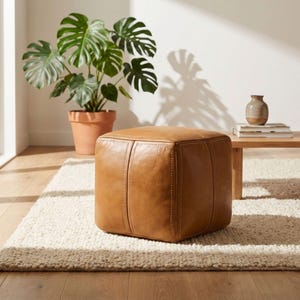 A square cognac tan genuine Moroccan leather pouf ottoman on a white rug in a bright boho living room with a green plant.