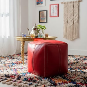 A vibrant red square Moroccan leather pouf ottoman sitting on a colorful bohemian rug next to a traditional Moroccan tea tray.