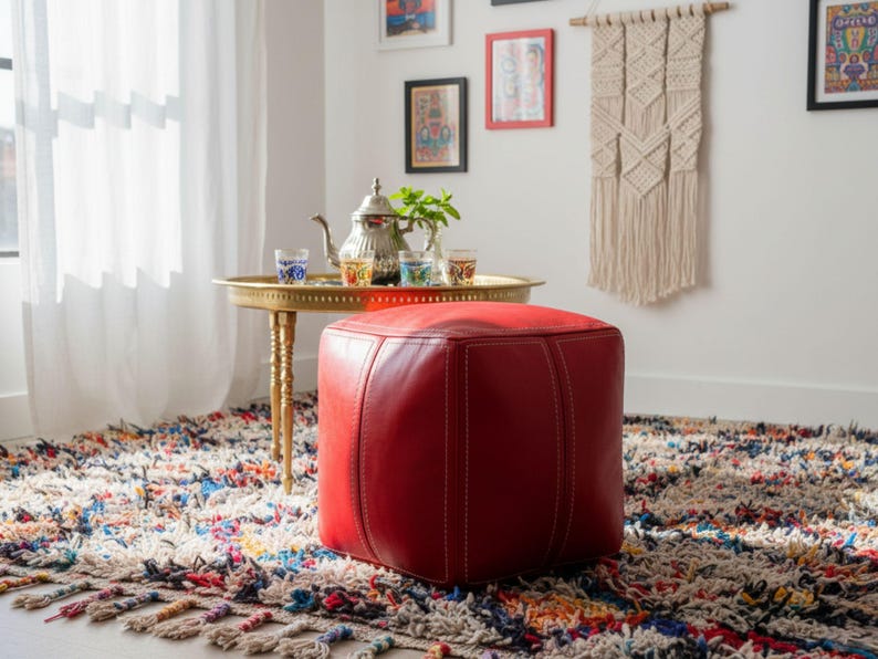 A vibrant red square Moroccan leather pouf ottoman sitting on a colorful bohemian rug next to a traditional Moroccan tea tray.