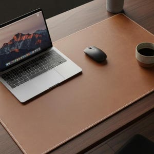 Könnte beinhalten: Eine braune Leder-Schreibtischunterlage mit weißen Nähten, ein Laptop, eine schwarze Maus und eine Kaffeetasse. Der Laptop zeigt eine Berglandschaft. Der Schreibtisch ist aus dunklem Holz.