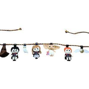 May include: A decorative garland featuring felt figures of characters and objects from a fantasy series. The garland includes a brown sorting hat, a figure in a black robe, a white owl, a figure with brown hair, a creature with large ears, a figure with red hair, a golden snitch, and a mandrake.